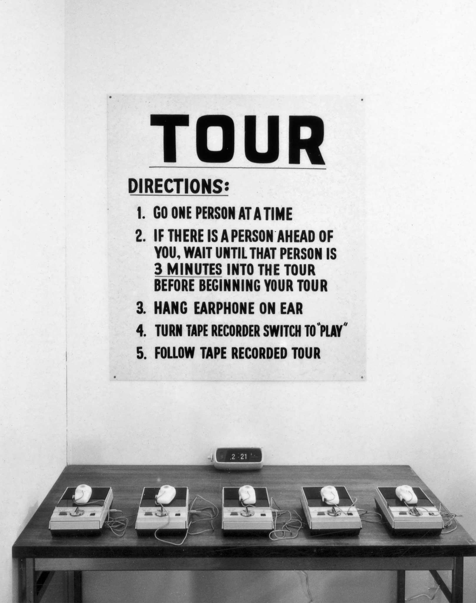 A table with five portable tape recorders and a large sign above it with directions for a guided audio tour